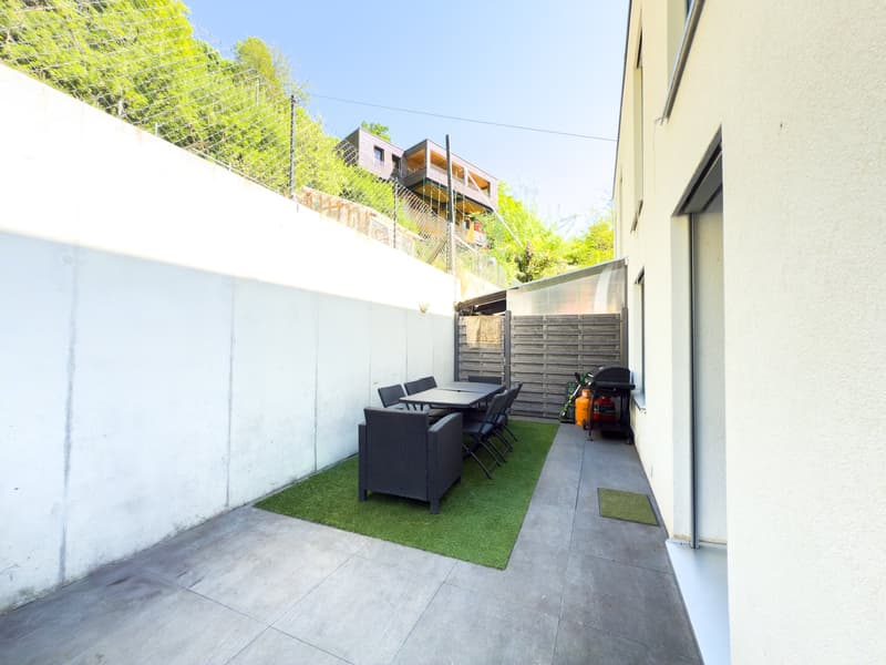 Paved patio area with artificial grass, outdoor furniture including a table and chairs, and a grill or barbecue. The space is surrounded by a fence and has a raised balcony or terrace above.