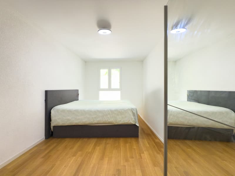 Spacious bedroom with hardwood flooring, white walls, and large windows. The room features a dark-colored bed frame and dresser, providing a modern and minimalist aesthetic.