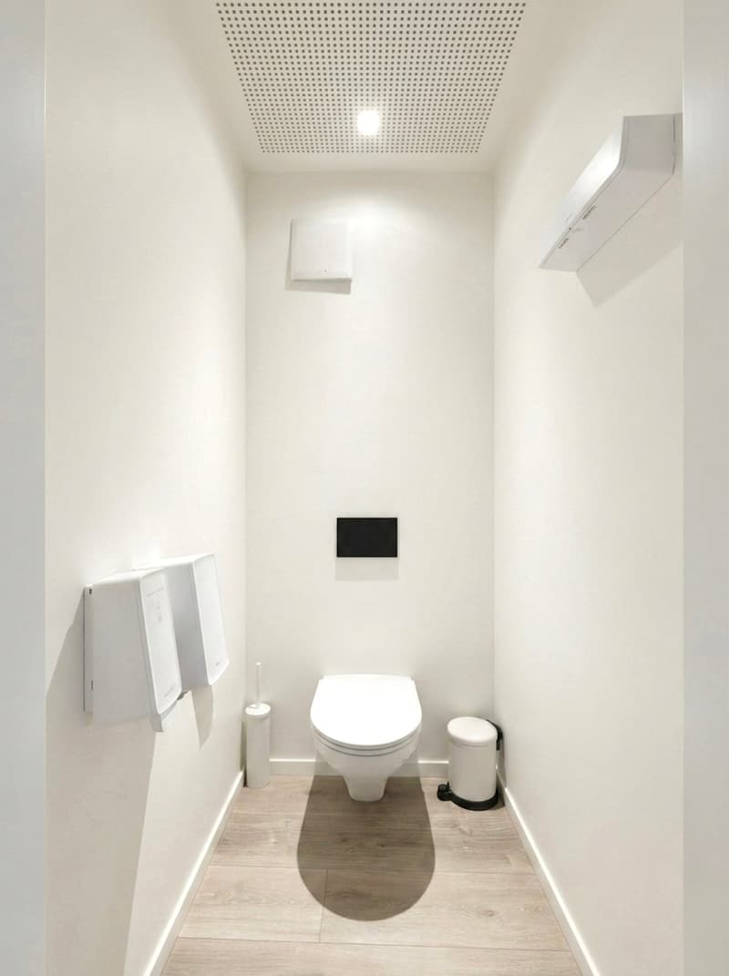 Small bathroom with white walls, wooden floor, a toilet, a trash can, towel dispensers, a ceiling light, and a vent.