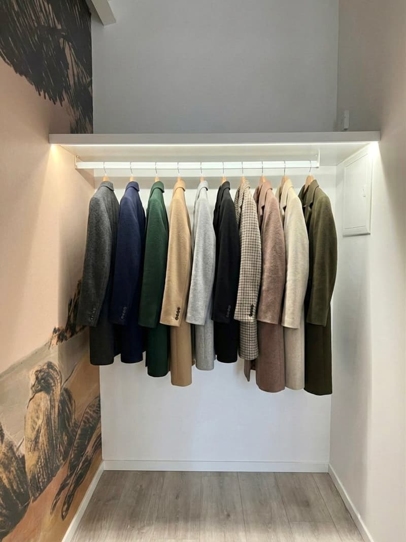 A room with a wall-mounted rack holding various coats. The wall is painted white, and there's a lit LED strip running along the ceiling of the rack. Below the rack, there is a wooden floor.