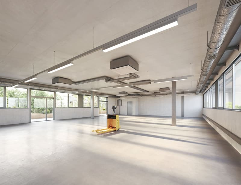 Large open commercial space with concrete floors, exposed ceiling, and overhead lighting. The space has large windows providing natural light and a forklift visible, indicating it may be used for storage or distribution purposes.