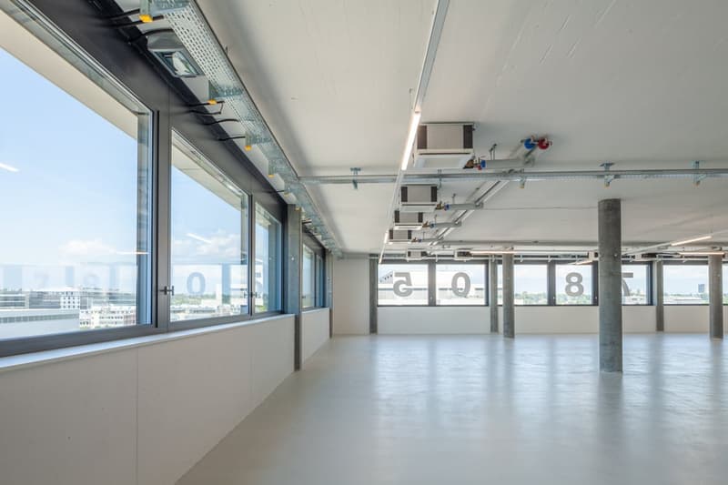 Large open office space with floor-to-ceiling windows, exposed ceiling with ventilation ducts, concrete columns, and numbered markings on the floor indicating different zones or sections of the space.