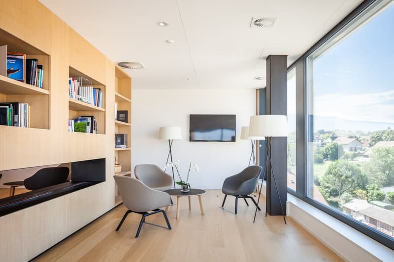Spacious living room with large windows providing a scenic view, wooden shelves with books, TV mounted on the wall, two gray armchairs, and a small coffee table