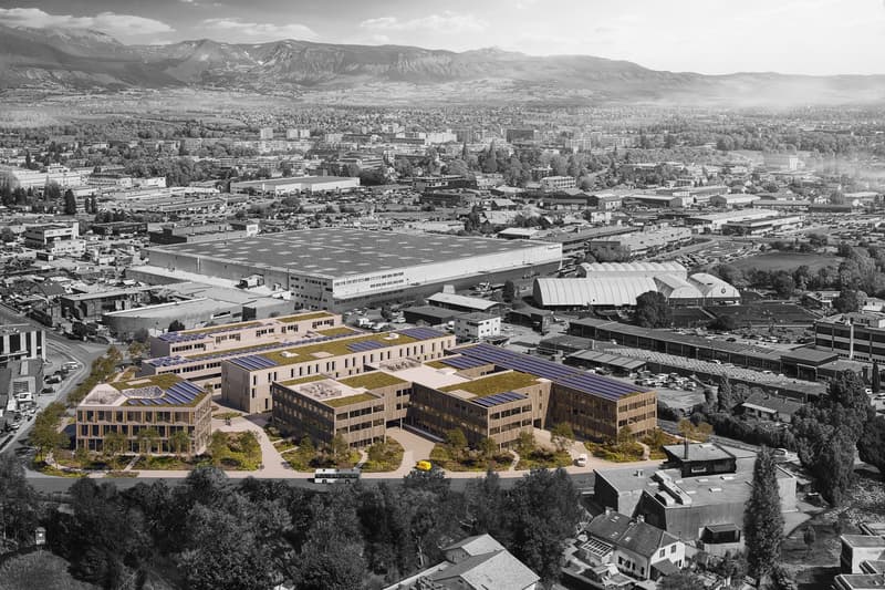 An office building with several solar panels on the roof, greenery around the building, mountains in the distance