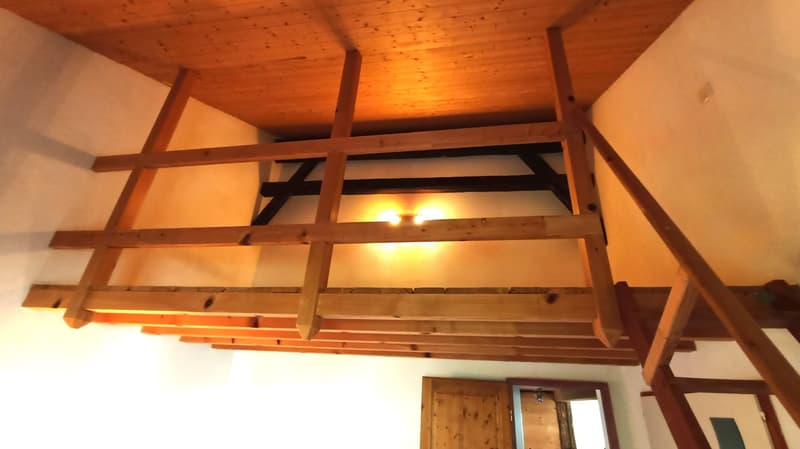 The image shows the interior of a room with a wooden ceiling and exposed wooden beams. The room has a warm, cozy atmosphere with the wooden elements and the lighting fixtures. There appears to be a doorway visible in the background.