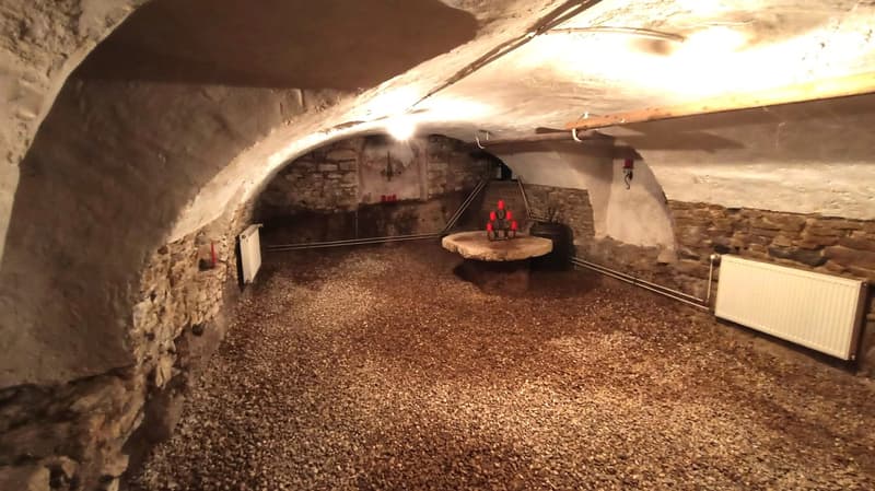 The image shows an underground space with a vaulted ceiling, stone walls, and a gravel floor. There is a wooden table with some objects on it, and what appears to be a heating or ventilation system visible on the ceiling. The space has a rustic, cave-like
