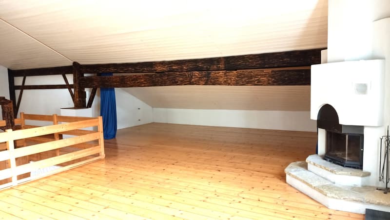 Large open space with wooden beams, wooden floor, and a fireplace. There are bunk beds or shelving units along one wall.