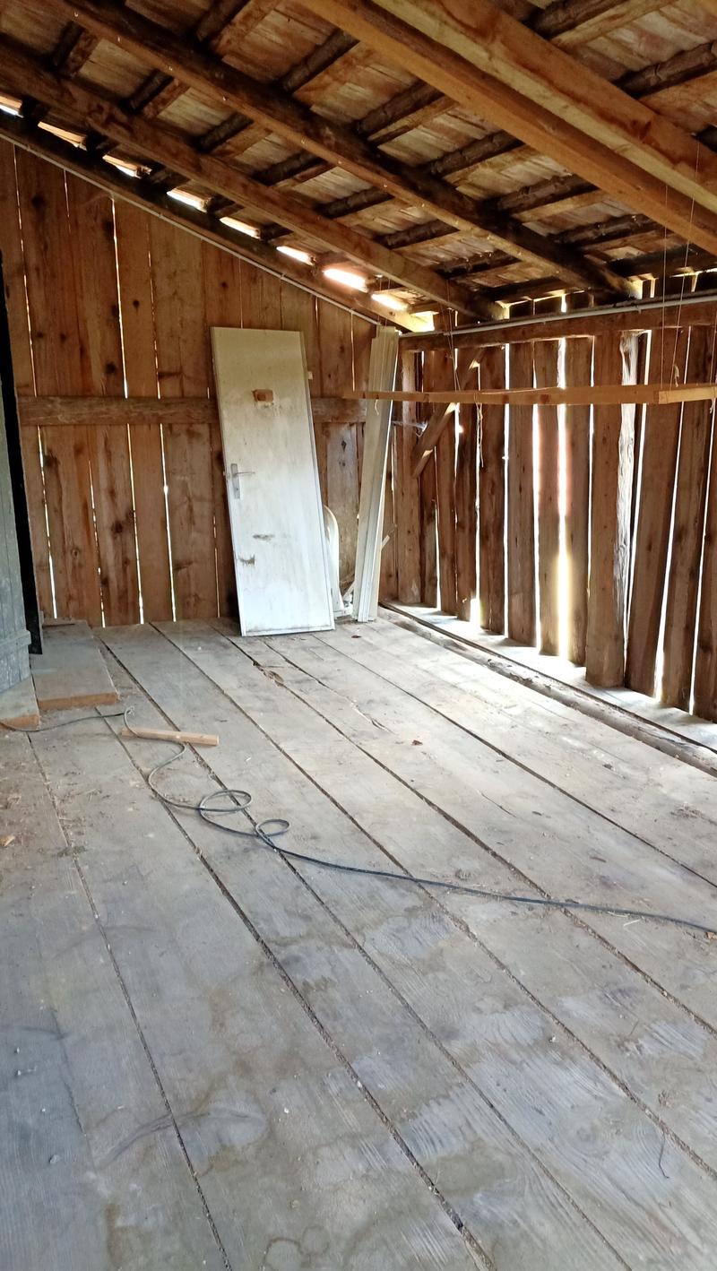 The image shows the interior of an old, wooden barn or shed. The walls are made of wooden planks, and the ceiling has exposed wooden beams. The floor is also made of weathered wooden planks. There is a white door visible in the corner of the room.