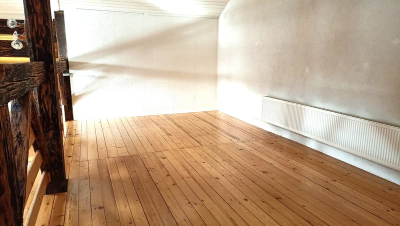 The image shows a spacious room with a wooden floor and white walls. The room appears to be empty, with no furniture or decor visible. The wooden floor boards are in good condition and run the length of the room. The walls are plain and unadorned, creatin