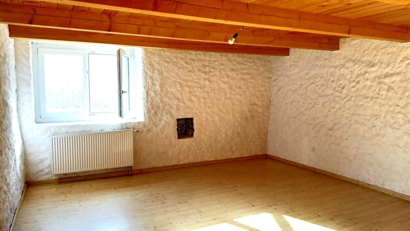 This image shows a room with a wooden ceiling, exposed wooden beams, and a large window. The walls are made of a textured material, possibly plaster or stucco. There is a radiator visible on one of the walls. The floor appears to be made of wood planks. T