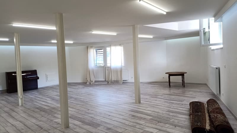 Empty room, wooden floor, white walls, central ceiling lights, white curtains on the window, piano, table, rolled up carpets.