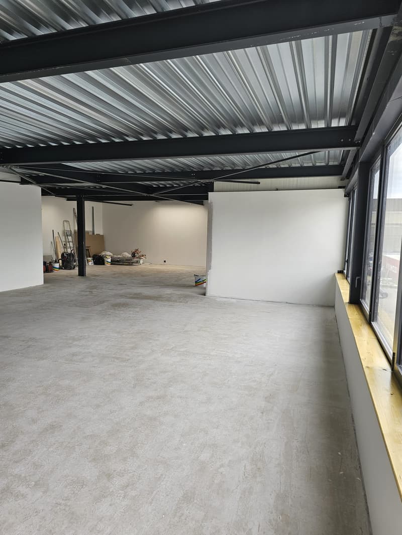 The image shows a large open space with a high ceiling and a metal frame structure. The floor is made of concrete and there are various items and equipment stored in the space. The walls are white and the overall atmosphere suggests this is a warehouse or