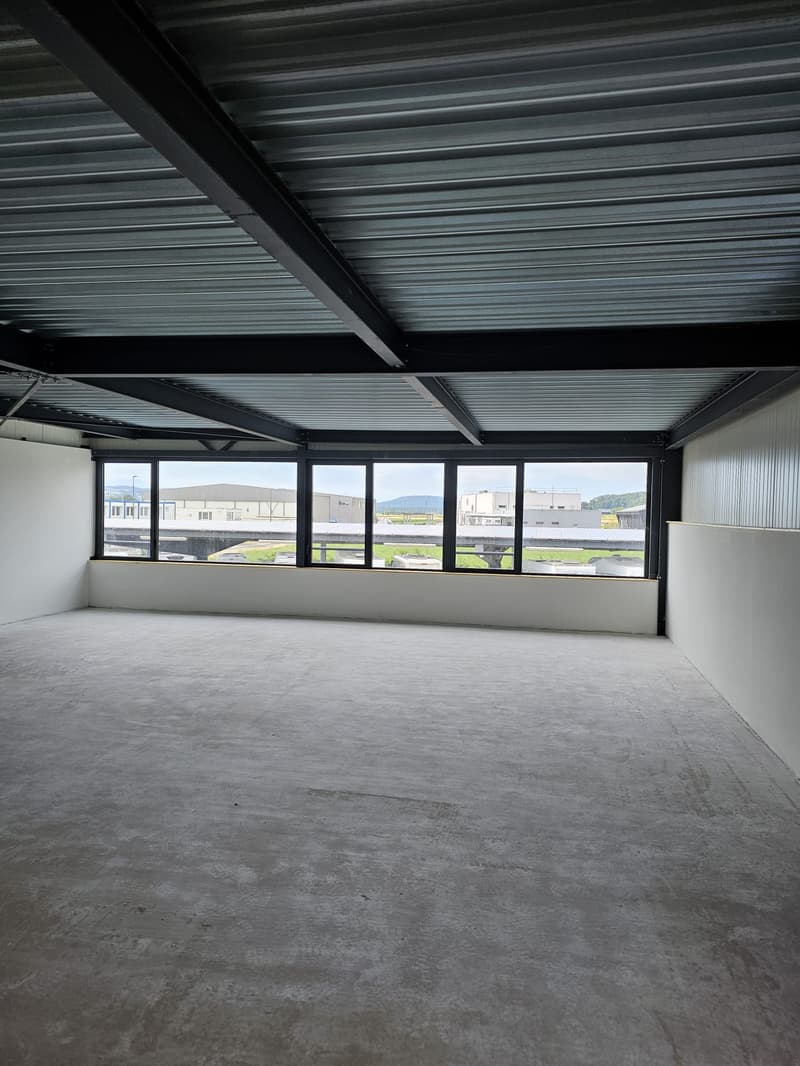 The image shows a large, open commercial space with a high ceiling and large windows that provide a view of the surrounding buildings and landscape. The space has a concrete floor and a metal roof structure with exposed beams. The overall impression is of