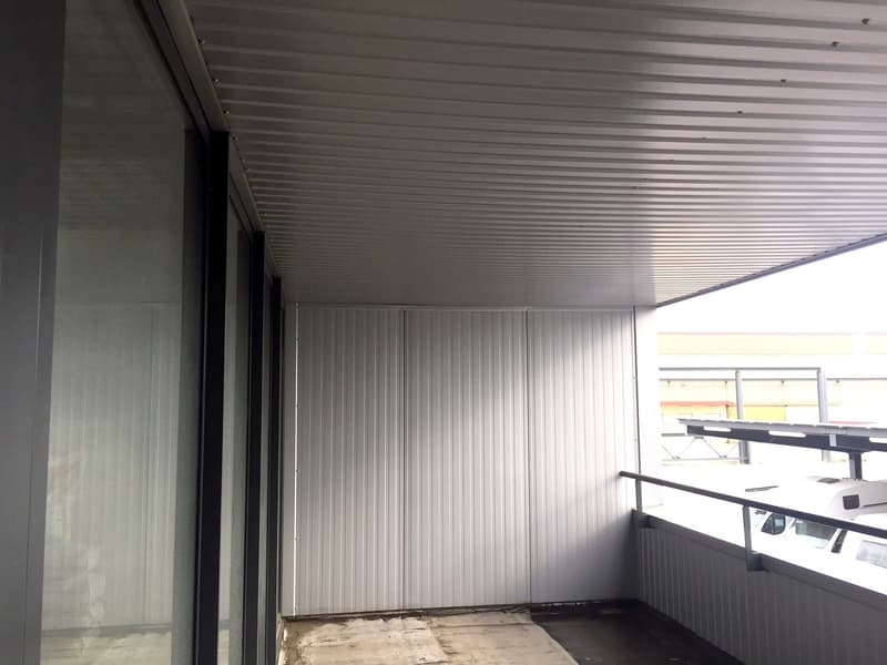 The image shows a room with a corrugated metal ceiling and walls. There is a glass window and a table or bench visible in the room.