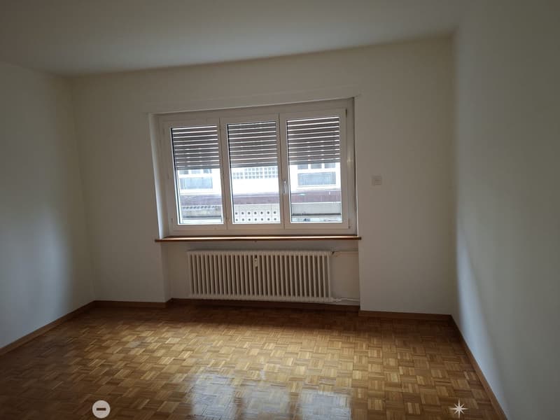 Empty room with large window, radiator, wooden floor, white walls.