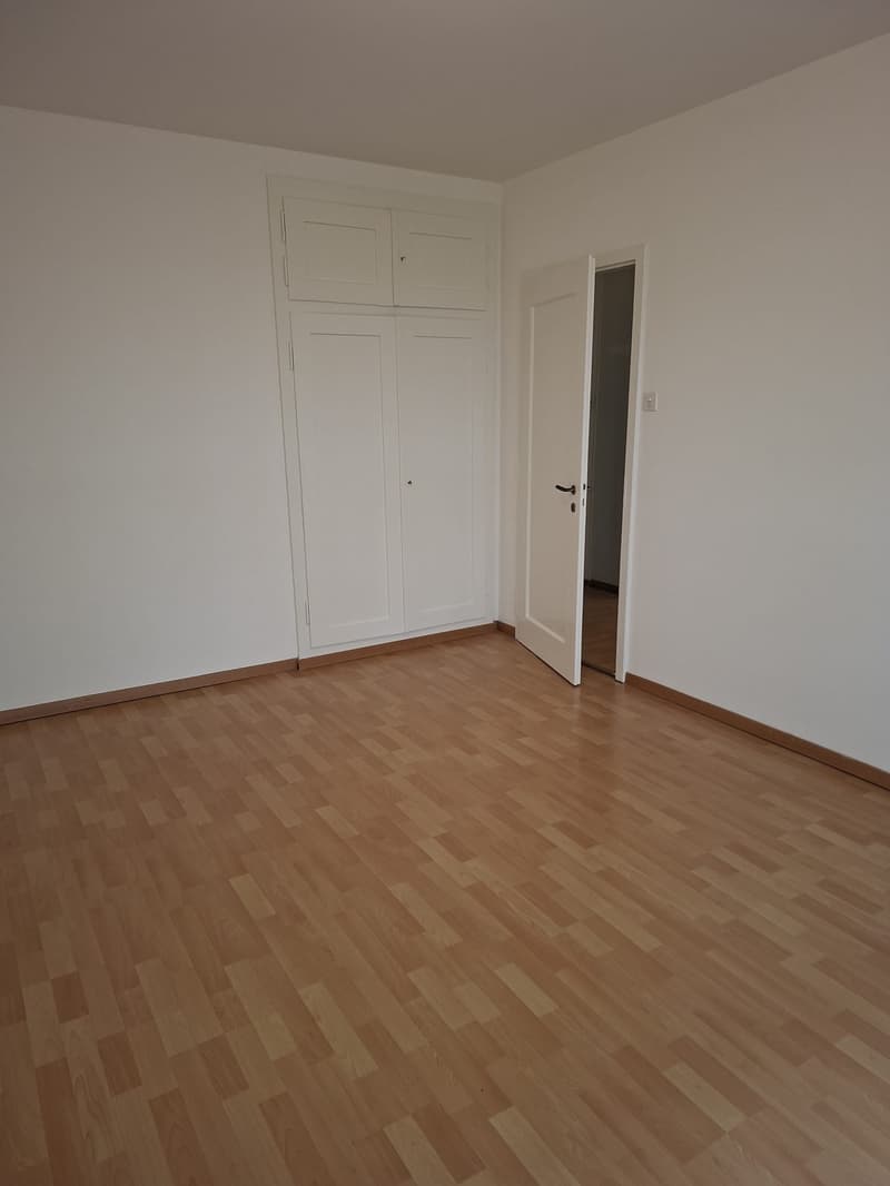 Empty room with white walls, wooden floor, and wooden cabinets. An open door leads to another room.