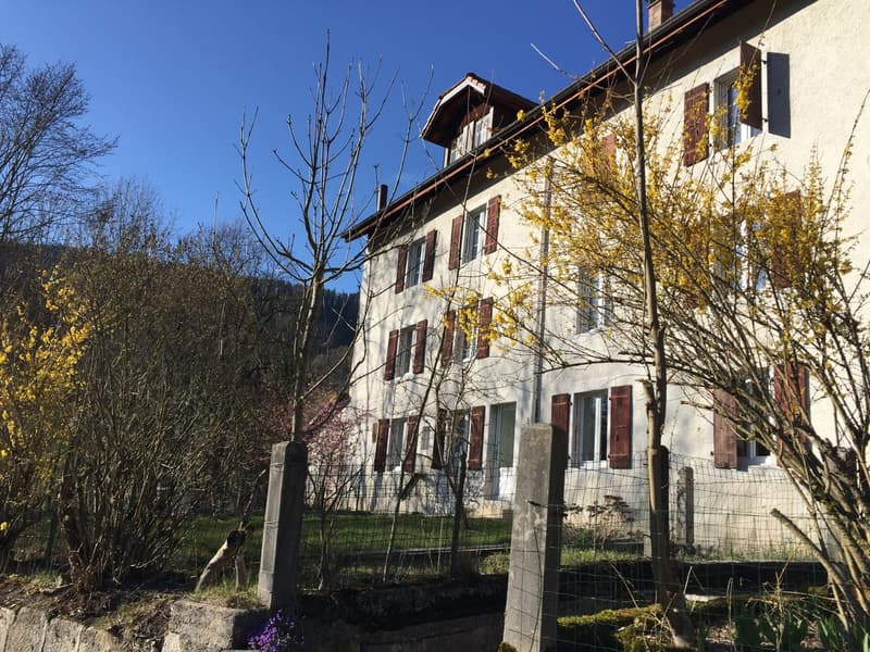white building with multiple windows, brown shutters, garden in front, mountains in the background