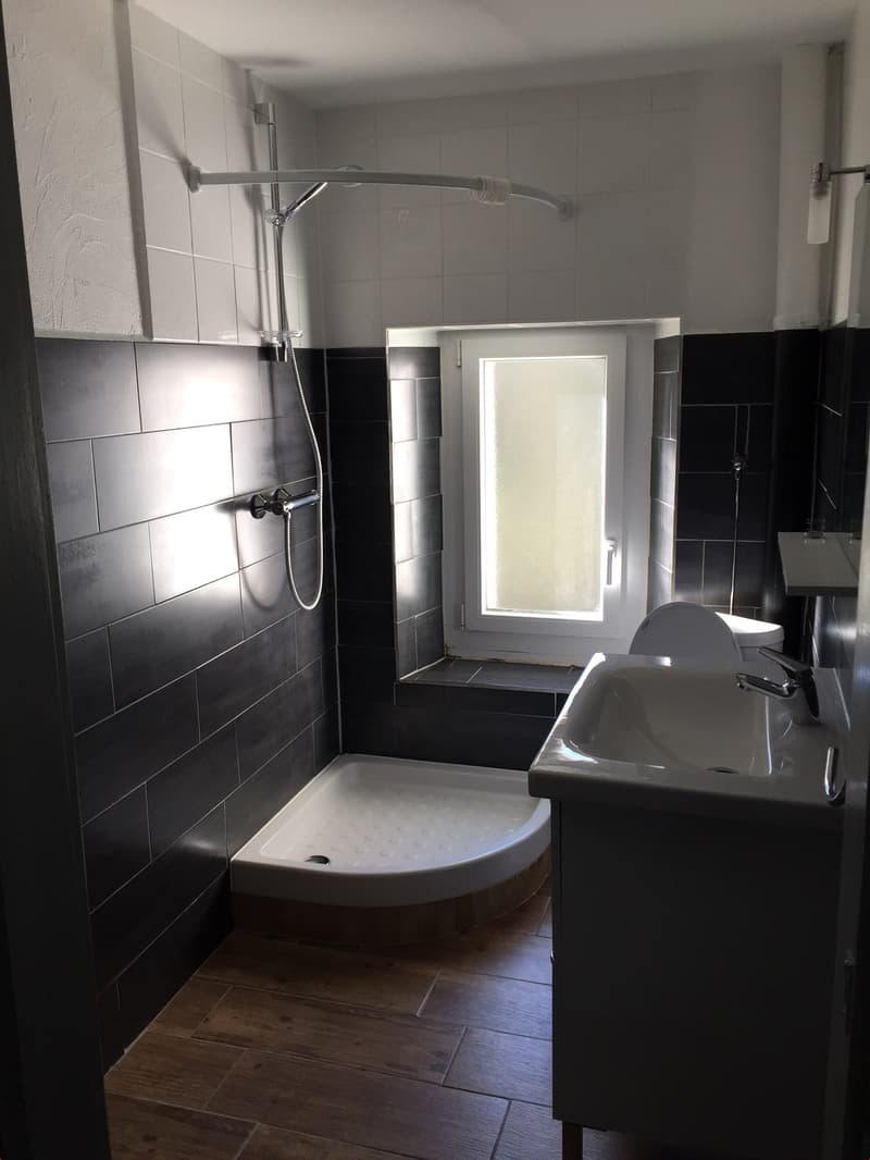 small bathroom with walk-in shower, black tiled walls, white sink, wooden floor