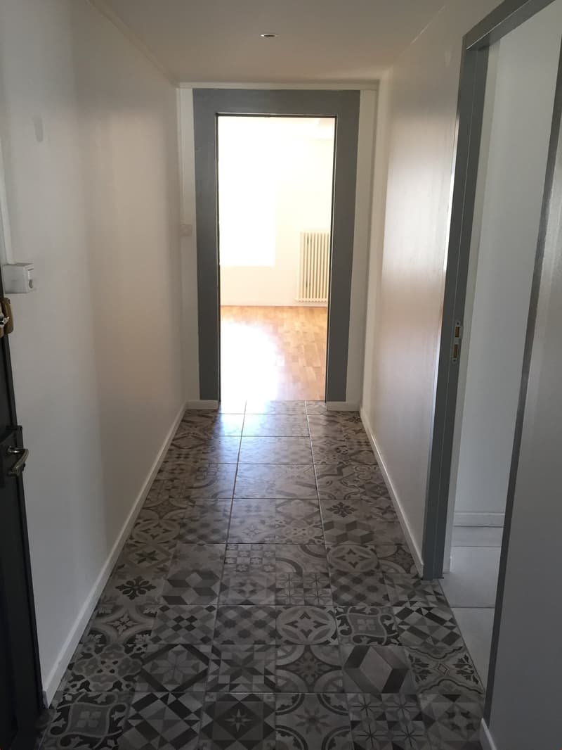 A hallway with gray walls, tiled floor, two doors, one leading to a white room with a wooden floor and white radiator.