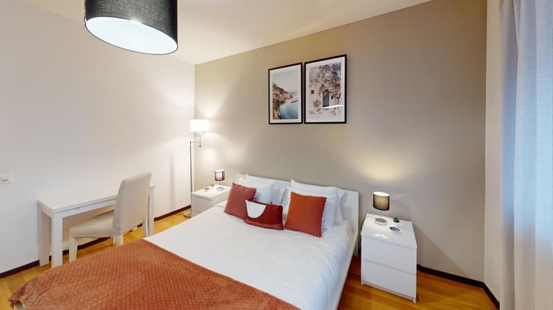 Bedroom: white walls, hardwood floor, bed with pillows, desk with chair, nightstands with lamps, two framed pictures on wall, white curtains