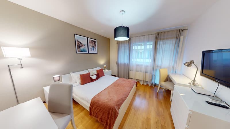 Single bed, white walls, wooden floor, large windows, lamps, night tables, framed paintings, television, chairs, desk, remote control