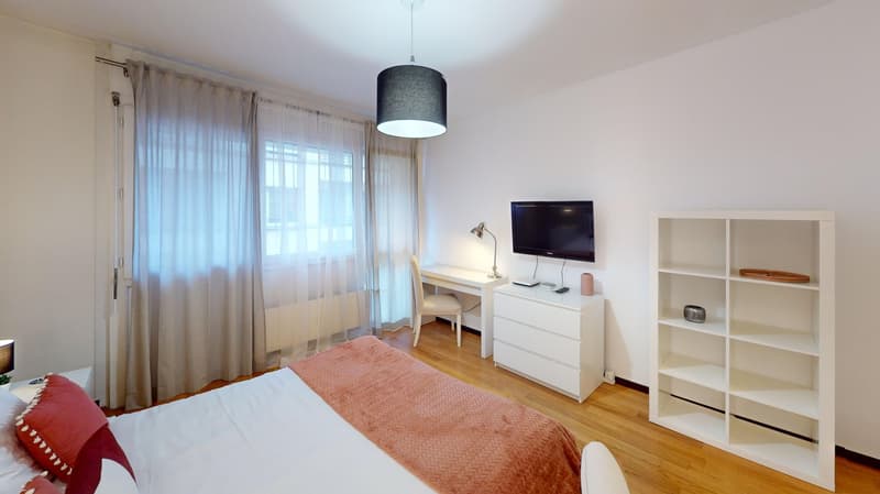 A bedroom featuring a white bed, white nightstand, desk with a chair, shelves with a book, small lamp, TV, and open curtains near a window.