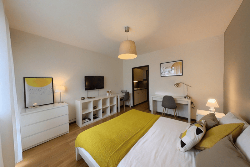 Spacious bedroom with white walls, hardwood floors, large windows, a white dresser, a desk, and a yellow bedspread. The room also features a TV mounted on the wall and a pendant light fixture.