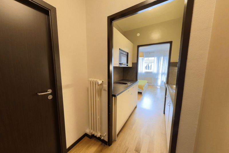 The image shows a hallway leading to a small kitchenette area. The hallway has wooden floors and beige walls. The kitchenette has a refrigerator, a microwave, and a dishwasher visible.