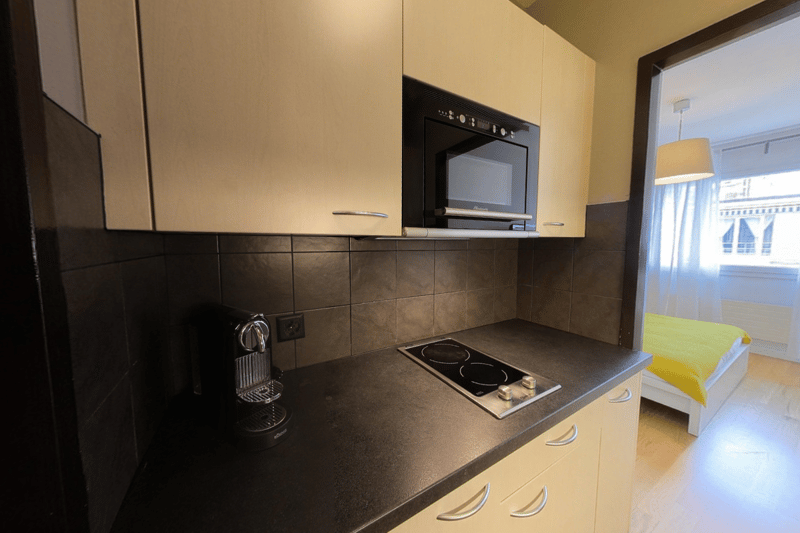 Compact kitchen with stove, microwave, coffee machine, countertop, cabinets, bed visible