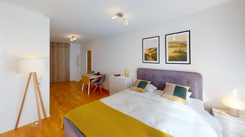 Bedroom with white walls, wooden floor, purple headboard, bed with white sheets and yellow blanket, white nightstands, two lamps, wooden floor, table with chairs, framed paintings on wall.