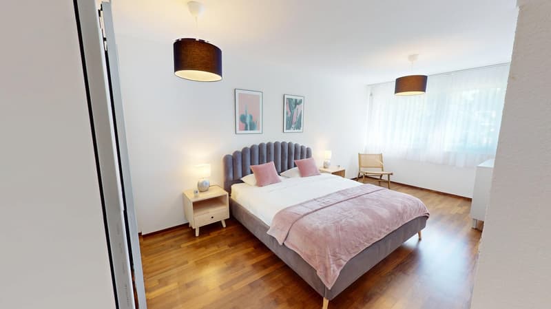 Wooden floor, white walls, large bed with pink blanket, two nightstands with lamps, two paintings on wall, wooden chair, white curtains