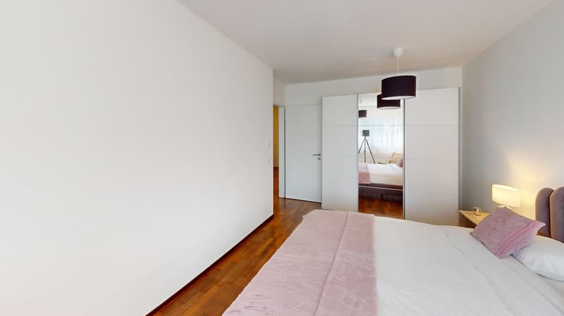 Bedroom with large bed, white walls, wooden floor, a nightstand, and a lamp
