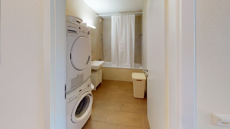 Laundry room with two stacked washers, white tiles, white walls, bathtub, sink, shower curtain, trash bin