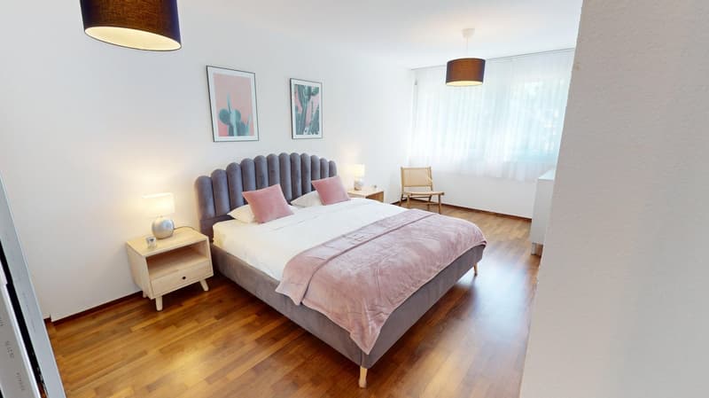 wooden floor, large double bed, gray headboard, pink bedding, two nightstands with lamps, beige armchair, framed pictures on wall, window with white curtains
