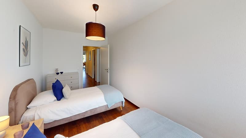 bedroom with wooden floors, white walls, two single beds, blue pillows, bedside table, white dresser, artwork on wall, open door
