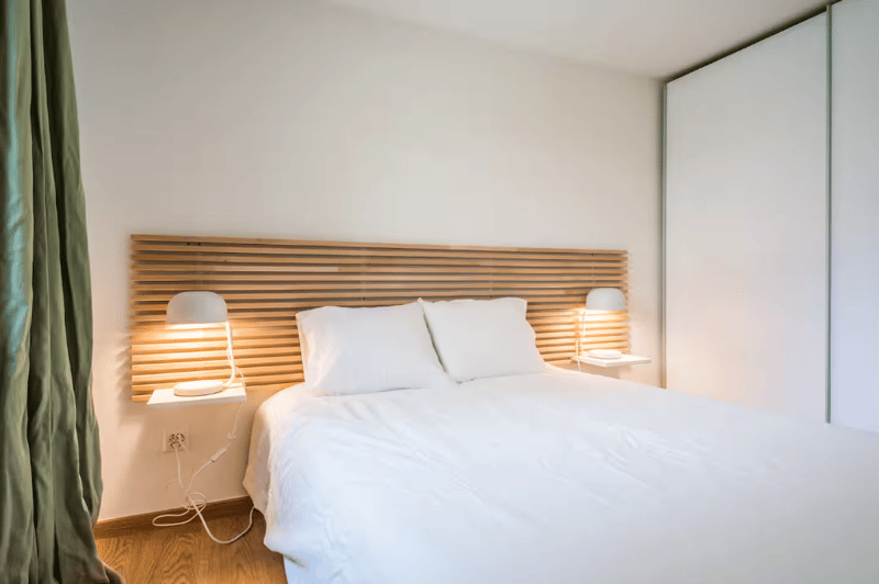 Queen size bed, wooden headboard, two lamps, two white pillows, power outlet, white bedding