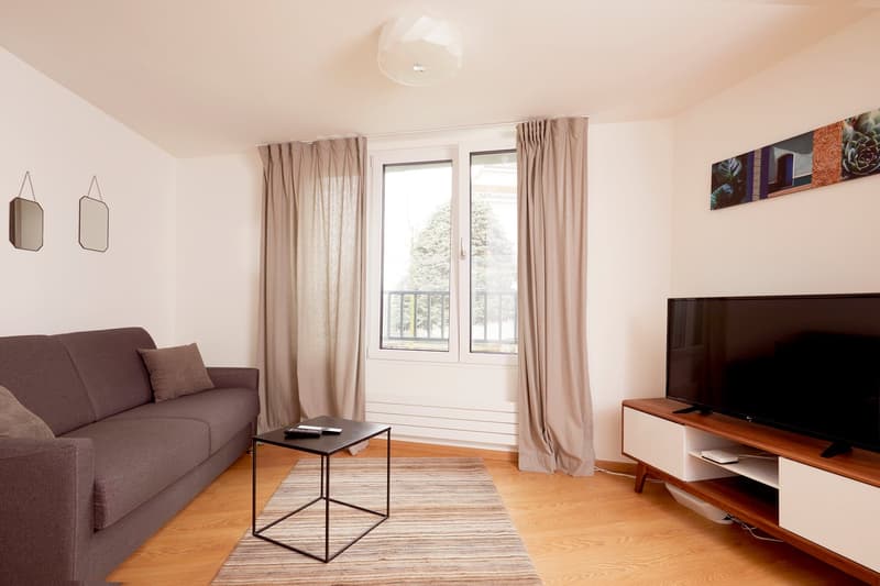 The image shows a cozy living room with a gray sofa, a wooden TV stand, and a patterned rug on the hardwood floor. The room has large windows that provide natural light and a view of the outdoors. The walls are painted in a neutral color, and there are de