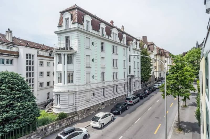 Multi-story building with a classical architectural style, featuring a white facade, ornate windows, and a red-tiled roof. The building is surrounded by trees and other residential buildings, with cars parked on the street in front.