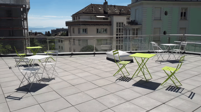 Outdoor terrace with tables, chairs, plants, buildings, mountains, sea, railing, sunlight, shadow