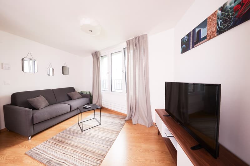 Spacious living room with large windows, gray sofa, coffee table, and TV stand. The room has hardwood floors and decorative mirrors on the wall.