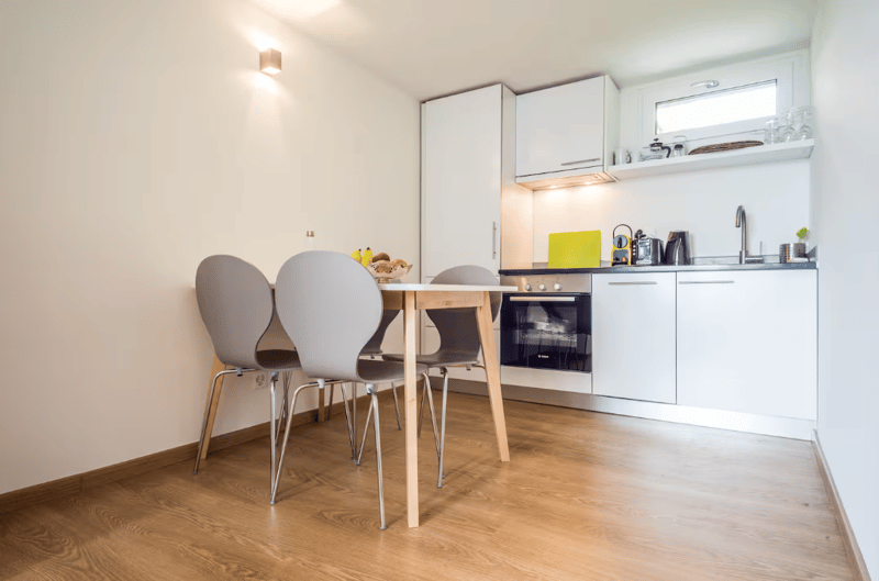 Kitchen with dining area, modern appliances, wooden floor, white cabinets, large windows, fruit bowl on table