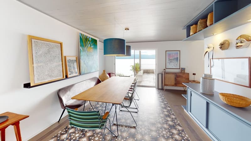 Dining room features a long wooden table, seating for several people, framed artworks on walls, patterned flooring, and a glass door leading to a view of the outdoors. A sideboard with a lamp is placed on one side.