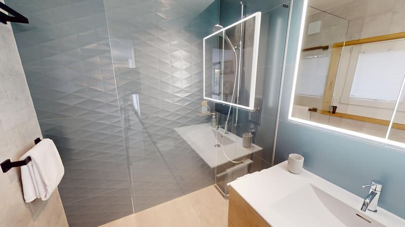 Modern bathroom with geometric tiled walls, shower area with frameless glass enclosure, a white vanity with a sink, a white towel bar, and a mirror with lit up sides.