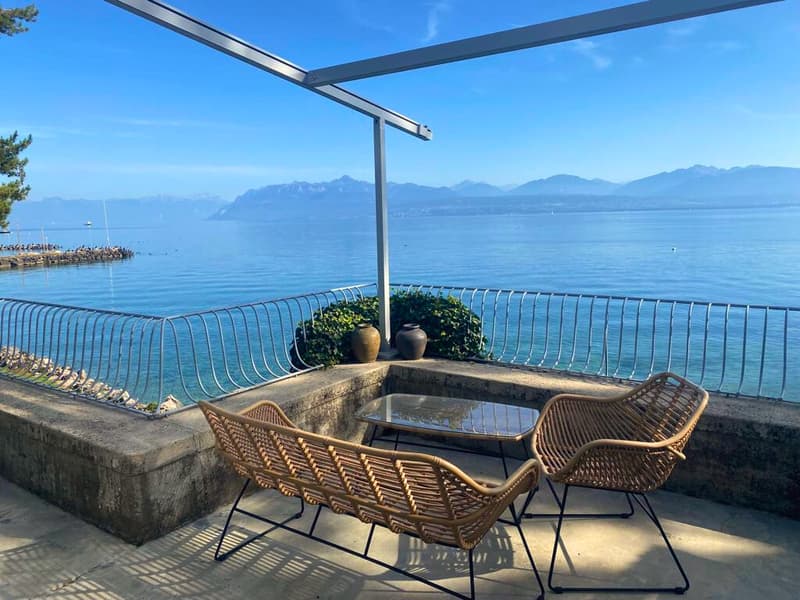 A balcony with a glass table and two wicker chairs, overlooking the sea with mountains in the distance