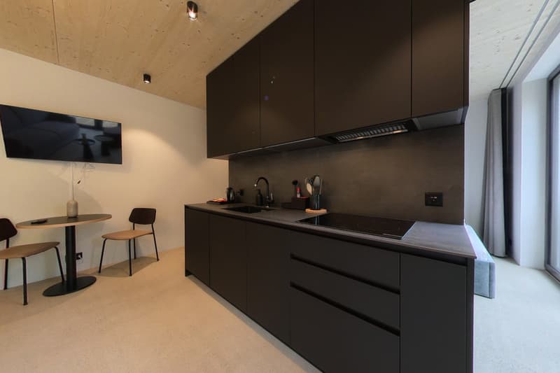 small modern kitchen, black countertops, black cabinets, black induction hob, 2 stools, 1 table, flat screen TV mounted on wall