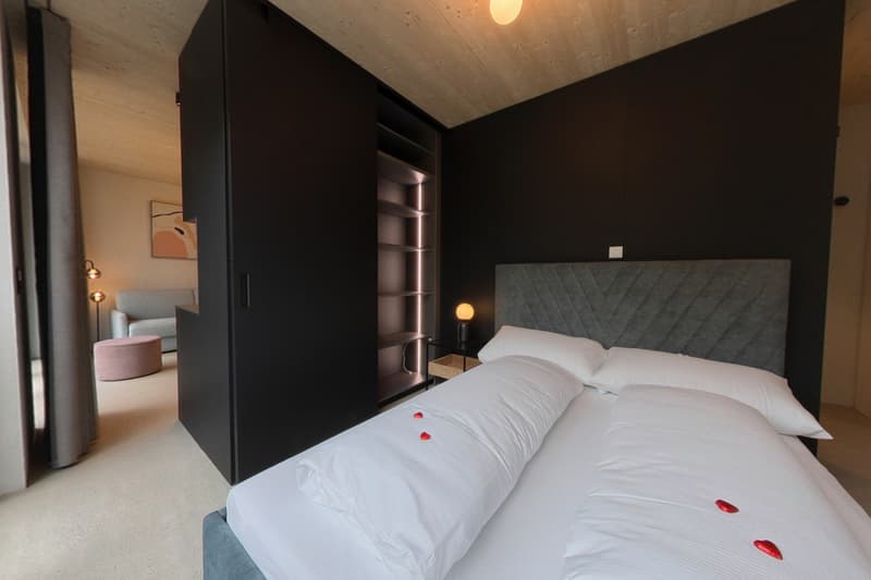 Large bed with pillows, gray headboard, wooden nightstand, lamp, black cabinets, hardwood floor, black walls, and open sliding door.
