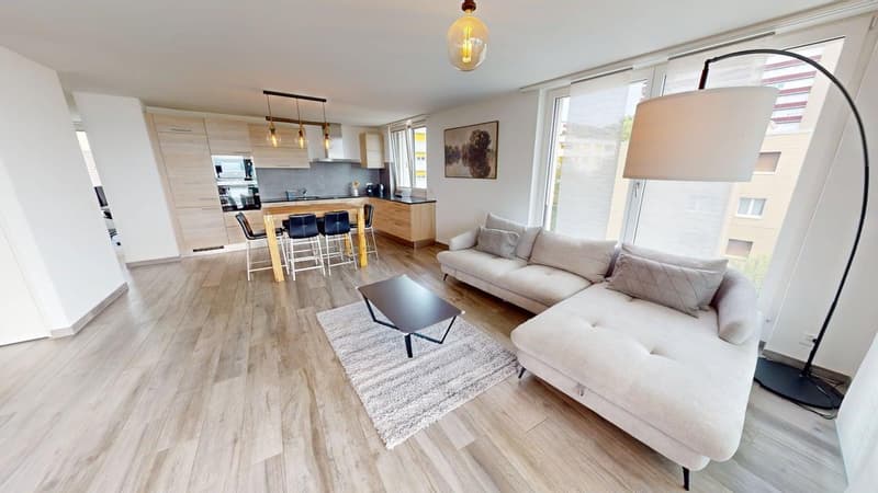 living room with L-shaped sofa, coffee table, rug, wooden flooring, and open to a kitchen with dining area, wooden table, chairs, modern kitchen cabinets, appliances, floor lamp, pendant lamps, large windows, and wall art