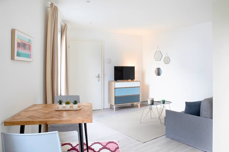 open floor plan, wooden table, small potted plants, gray sofa, wooden side table, small rug, picture frame, blue chevron chest of drawers, TV, small curtains, white walls