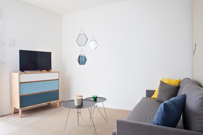 Interior of a modern living room with a comfortable couch and a wooden sideboard. The room features light-colored walls, a flat-screen TV on top of the sideboard, and two decorative hexagon mirrors on the wall. The carpet adds warmth to the room, and the