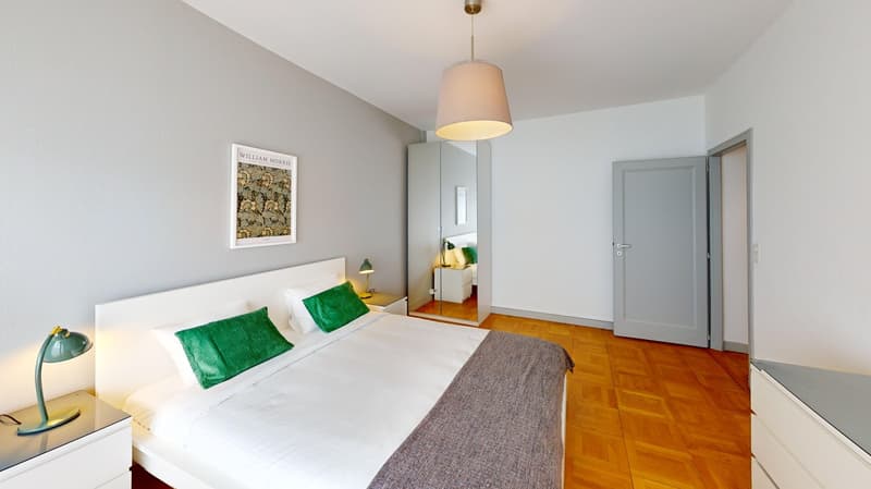 Bedroom with white walls, wooden floor, large bed, nightstands, lamps, mirror, picture frame, and a door.